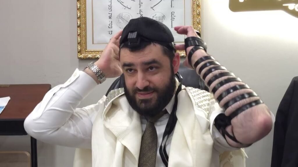 Bearded man in white prayer shawl adjusting tefillin on his head and arm during Jewish prayer.