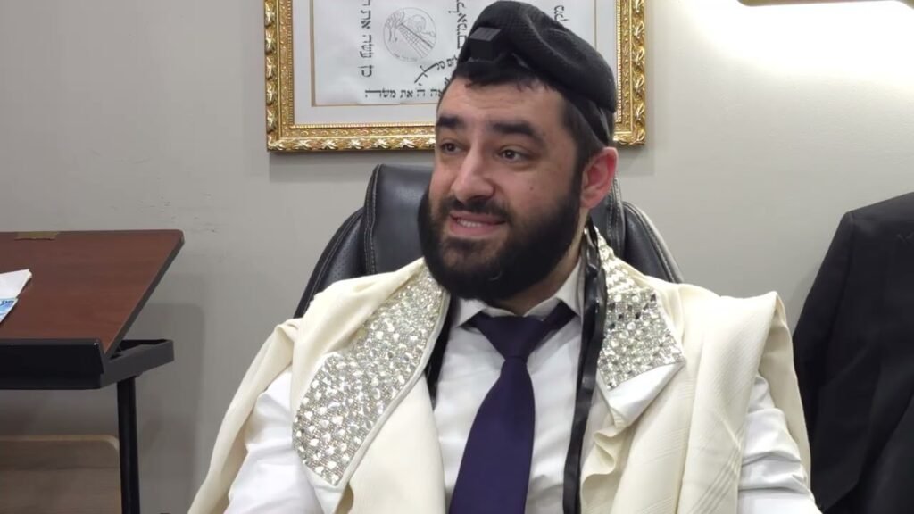Bearded man in a cream ceremonial suit with rhinestone-embellished lapels, a purple tie, and a head-mounted camera, seated in an office chair with a framed artwork behind him.