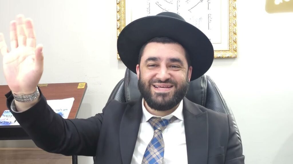Man in a dark suit and wide-brimmed hat sits at a desk in an office, smiling and waving at the camera.
