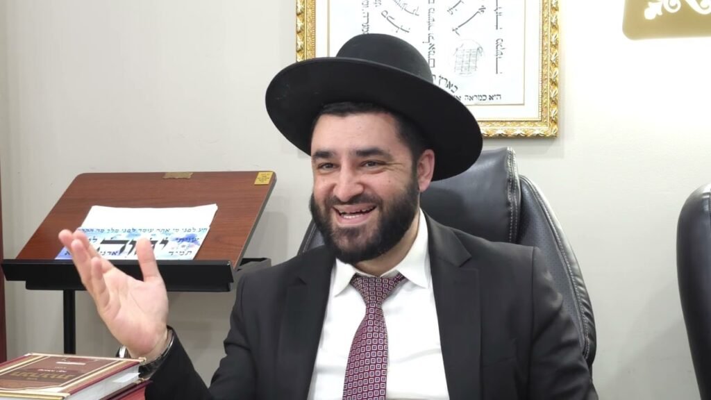 Bearded man in a black suit and wide-brimmed hat sits at a desk, smiling and waving his hand.