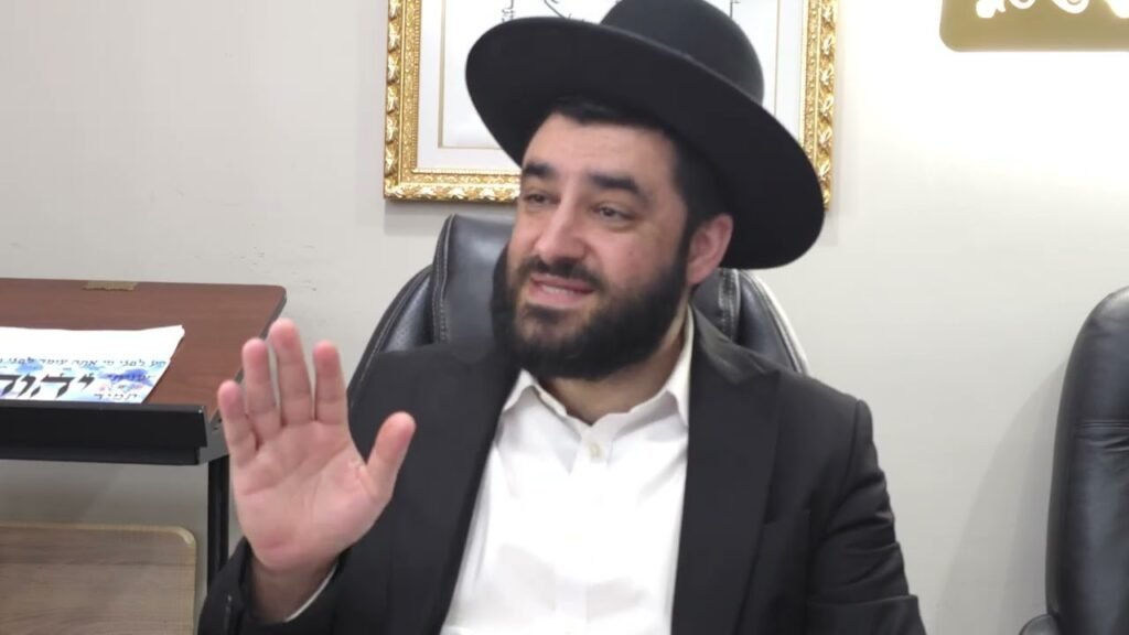 Bearded man in a black hat and suit raises his hand in a greeting indoors, seated in an office.