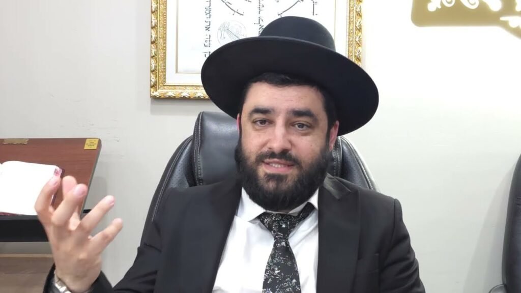 Bearded man in a black hat and suit sits at a desk, gesturing with his left hand during a discussion in an office setting.