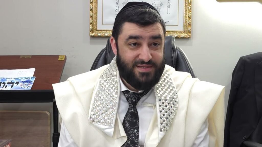 Bearded man in a white ceremonial robe and kippah sits in an office chair, smiling slightly.