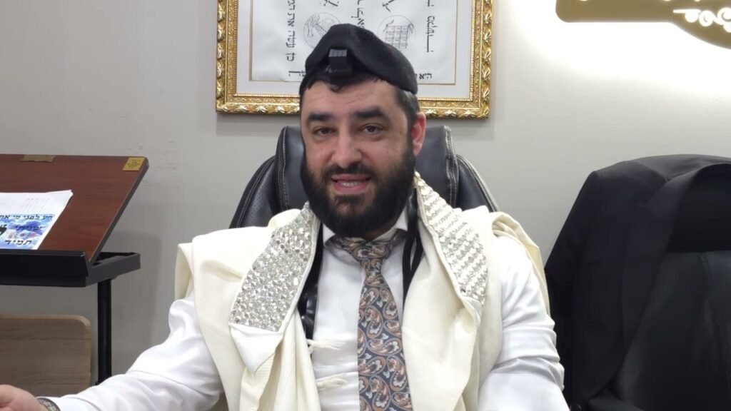 Bearded man in a ceremonial white robe and patterned tie sits at a desk in an office, wearing a kippah with a small device attached.